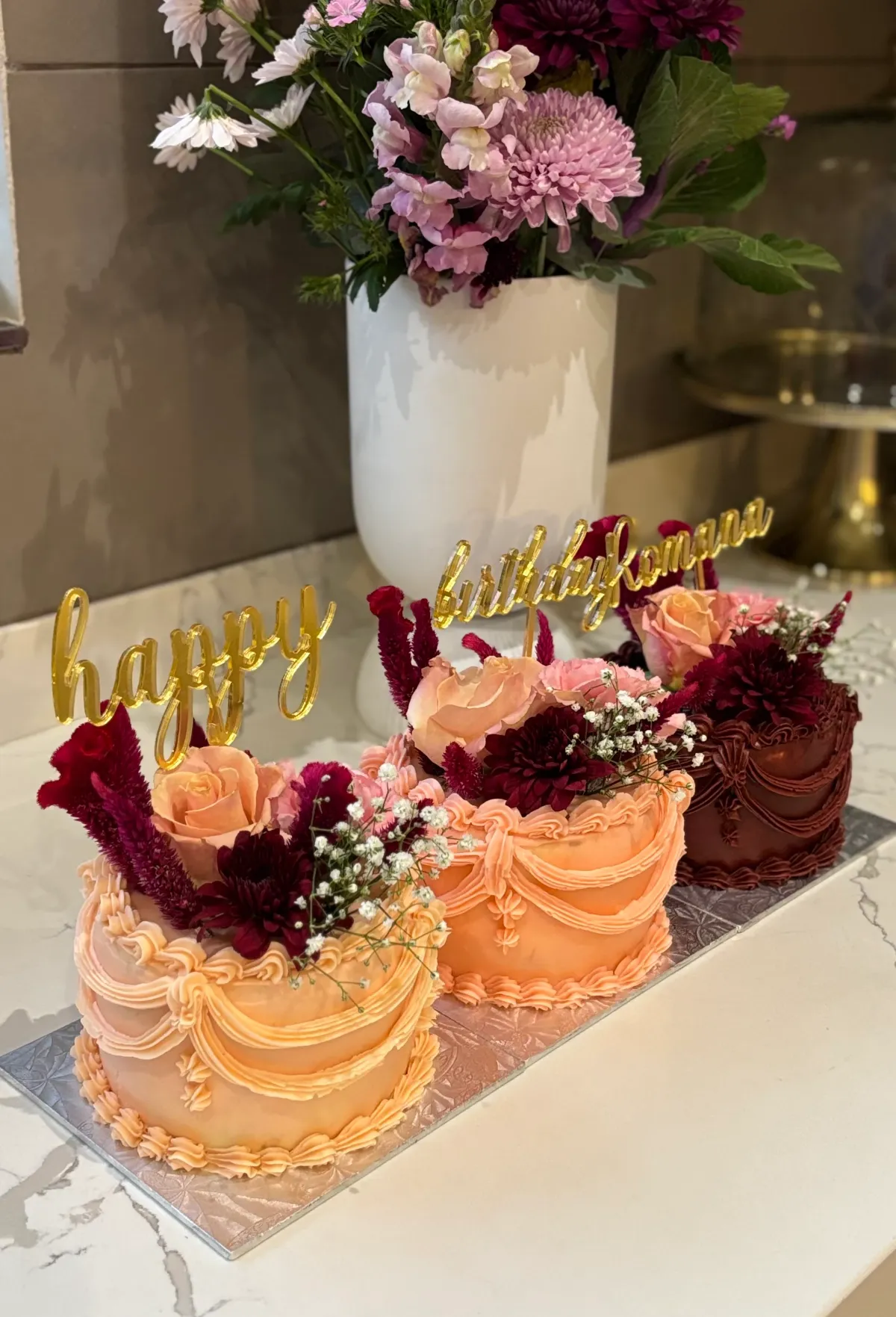 Three custom celebration cakes with peach and burgundy piped swags, fresh roses and dahlias, and gold script cake toppers