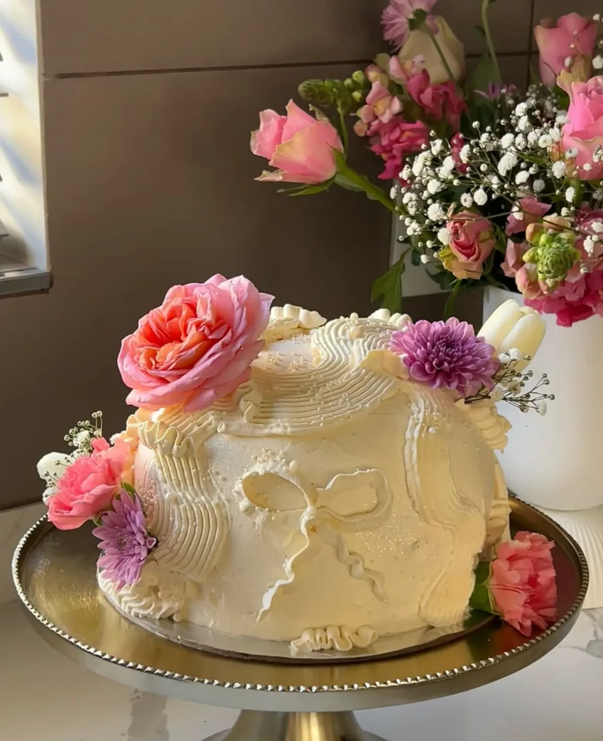 A white buttercream cake with a piped bow design, topped with fresh pink roses and purple chrysanthemums