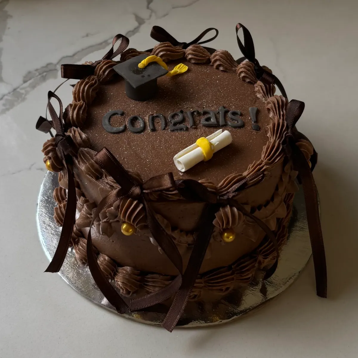 A chocolate graduation cake with a fondant cap and diploma, finished with brown ribbons and 'Congrats!' script