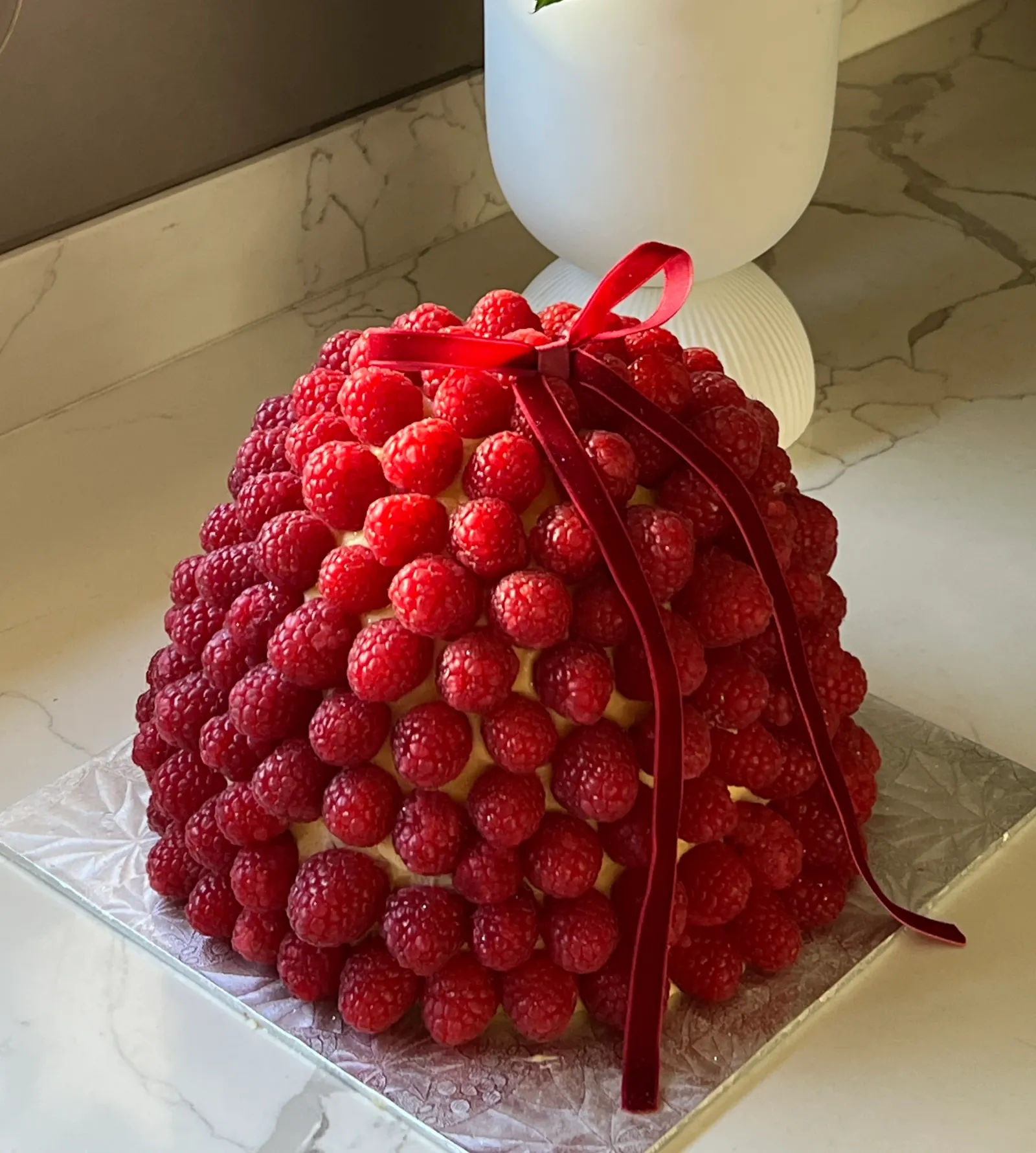 A dome cake covered in fresh raspberries, finished with a deep red velvet ribbon bow